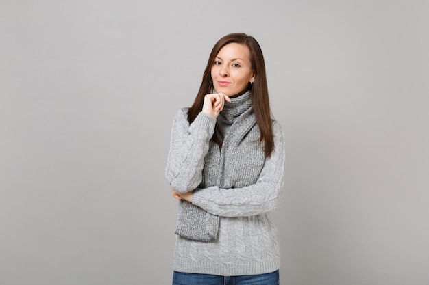 Young woman in gray sweater scarf put hand prop up on chin isolated on grey wall background, studio portrait. Healthy fashion lifestyle people sincere emotions cold season concept. Mock up copy space.