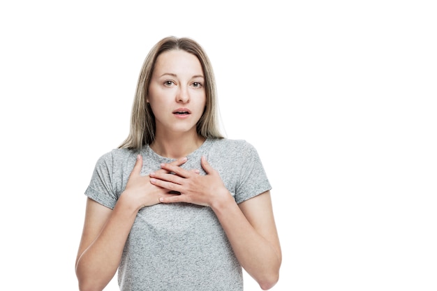 A young woman in a gray T-shirt is surprised to open her mouth and clasp her hands to her chest. Sincere emotion. Isolated on a white wall.