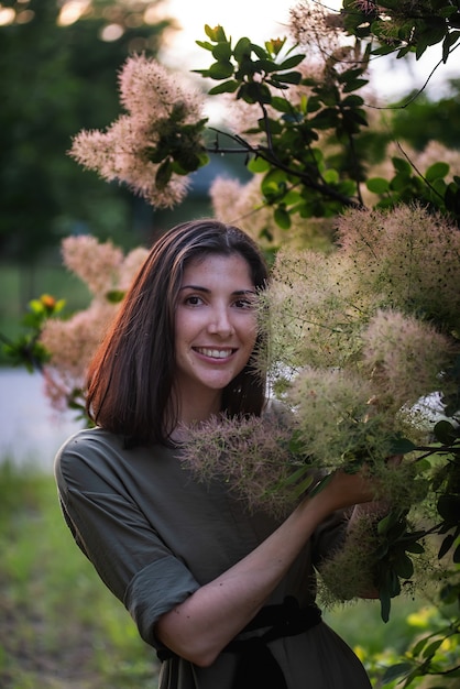 A young woman in a green khaki dress stands by a rosebush Smoky skumpia at sunset in the park.