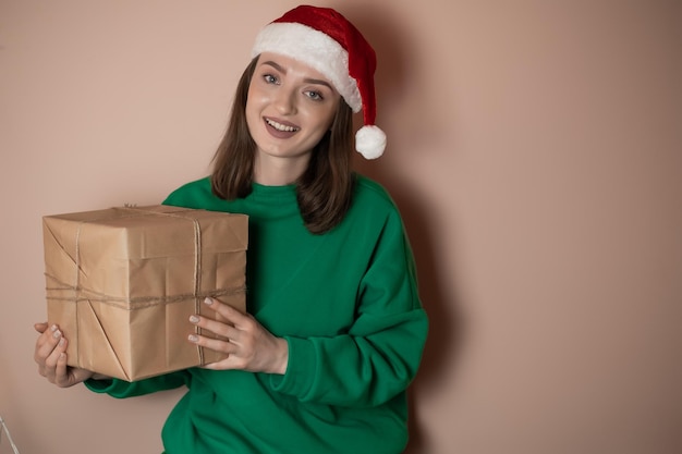 Young woman in a green sweater and in a New Year's hat holds a gift box on a simple green background Holiday concept Happy New Year 2023 chrome key merry christmas