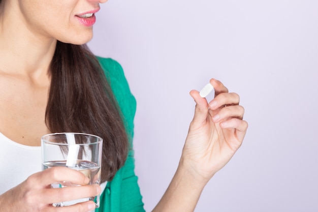 Young woman in green sweater and white shirt holding a pill and a glass of water. Medicine and health care concept.