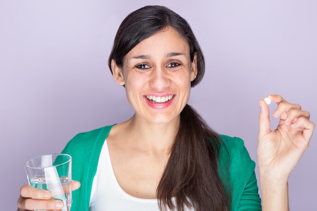 Young woman in green sweater and white shirt holding a pill and a glass of water, smiling looking at camera. Medicine and health care concept.