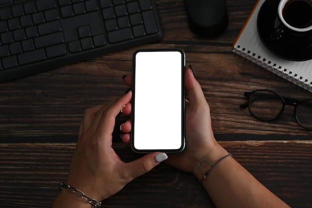 Young woman hands holding mockup smart phone with white empty screen at wooden desk