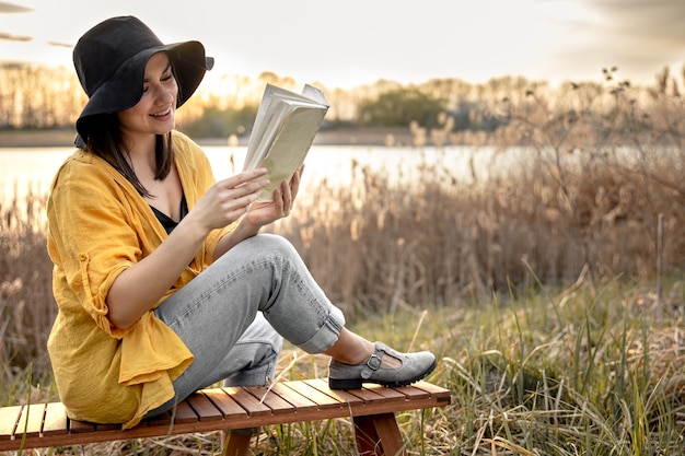 A young woman in a hat with a smile on her face is reading a book sitting by the river at sunset