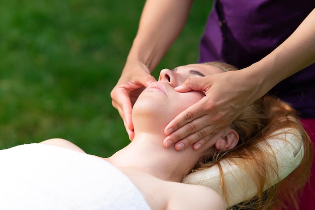 Young woman head getting face massage with doctor hands outdoors on natural background, enjoying, relaxing. Pretty female relaxed patient doing beauty treatment, healthy skin, anti age procedure.