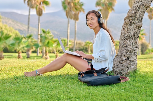 Young woman in headphones with laptop sitting on the grass in the park