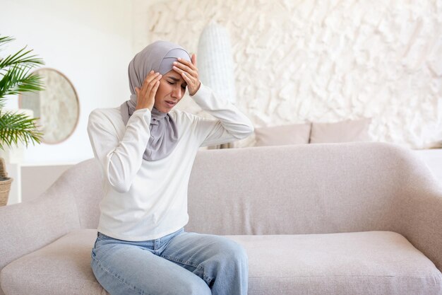 A young woman in a hijab is sitting on the sofa at home and holding her head with her hands feeling