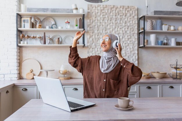 A young woman in hijab of oriental origin is sitting at home in the kitchen using a laptop and