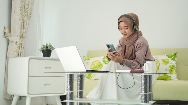 Young woman in hijab working from home looking for ideas on smartphone while listening on headphones