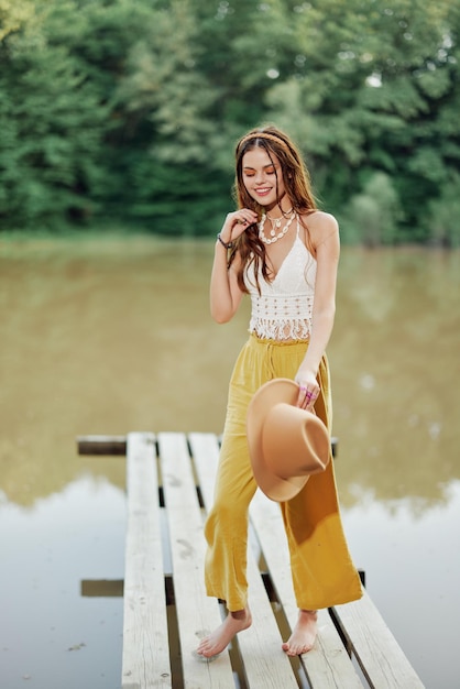 A young woman in a hippie look and ecodress dancing outdoors by the lake wearing a hat and yellow pants in the summer sunset