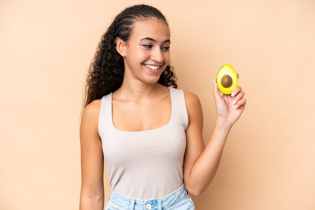 Young woman holding an avocado isolated on beige background with happy expression