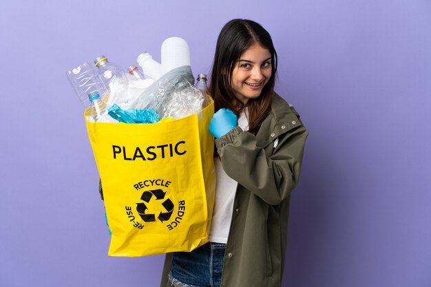 Young woman holding a bag full of plastic bottles to recycle isolated on purple background celebrating a victory