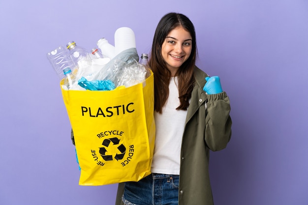 Young woman holding a bag full of plastic bottles to recycle isolated on purple celebrating a victory in winner position