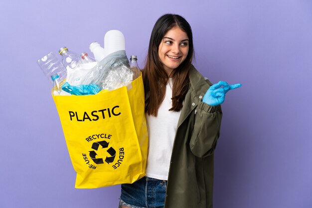 Young woman holding a bag full of plastic bottles to recycle isolated on purple wall intending to realizes the solution while lifting a finger up