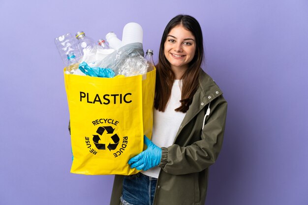 Young woman holding a bag full of plastic bottles to recycle isolated on purple wall looking to the side and smiling