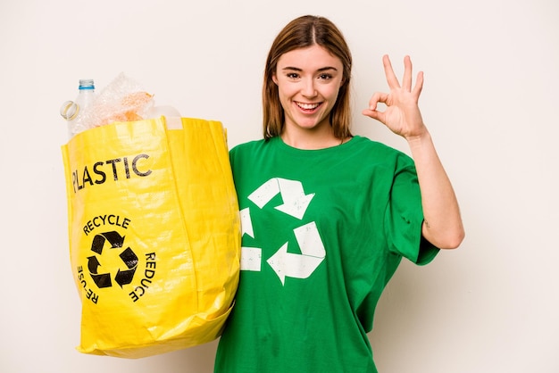 Young woman holding a bag full of plastic bottles to recycle isolated on white background cheerful and confident showing ok gesture