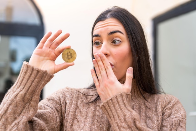 Young woman holding a Bitcoin at outdoors with surprise and shocked facial expression