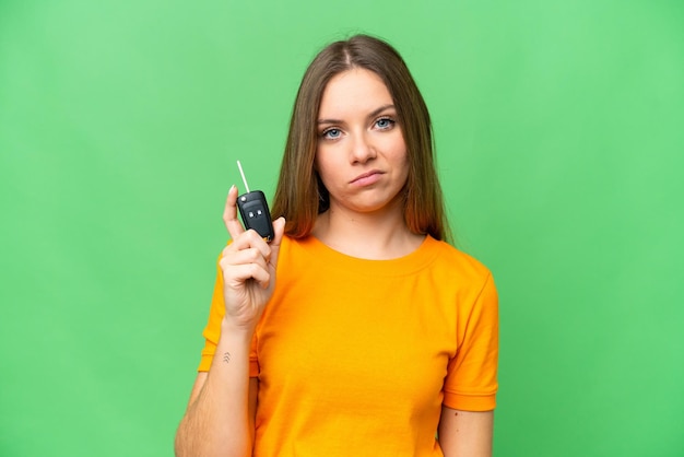 Young woman holding car keys over isolated chroma key background with sad expression