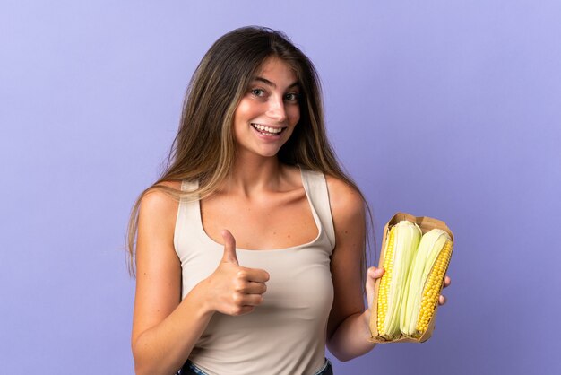 Young woman holding corn isolated on purple wall with thumbs up because something good has happened