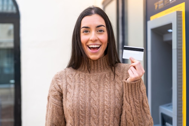 Young woman holding a credit card at outdoors with surprise and shocked facial expression