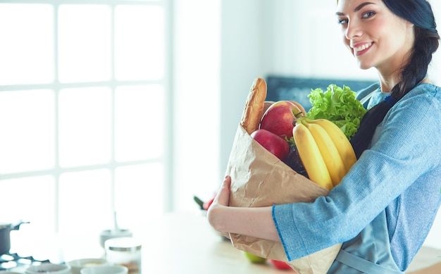 Young woman holding grocery shopping bag with vegetables standing in the kitchen