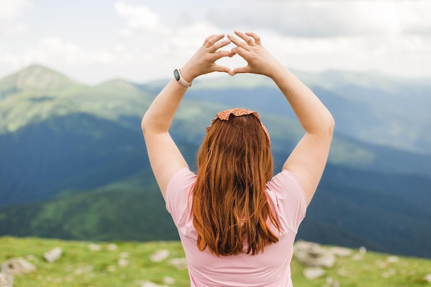 Young woman holding her hands up in the shape of heart against the background of the mountain valley Happy girl standing backwards looking away Traveling in the mountains Freedom travel concept