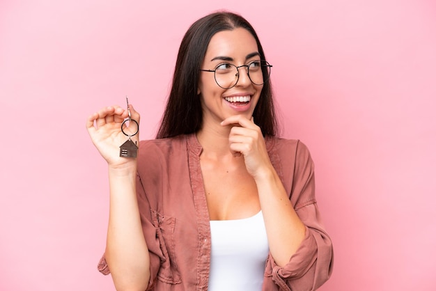 Young woman holding home keys isolated on pink background thinking an idea and looking side