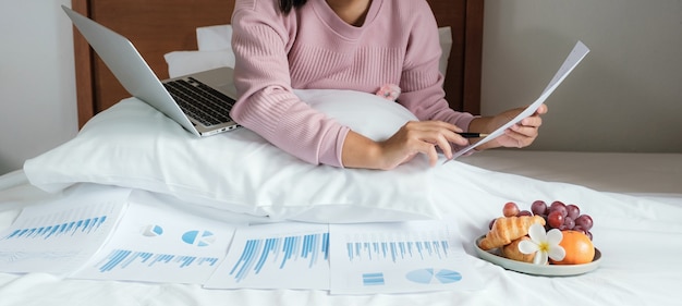 Young woman holding pen used laptop video conferences and eating fruits on the bed work from home