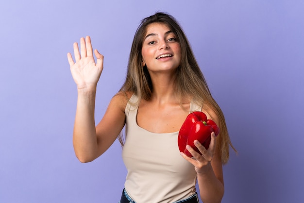 Young woman holding a pepper isolated on purple saluting with hand with happy expression