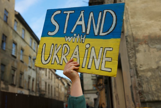 Young woman holding poster in colors of national flag and words Stand with Ukraine outdoors closeup