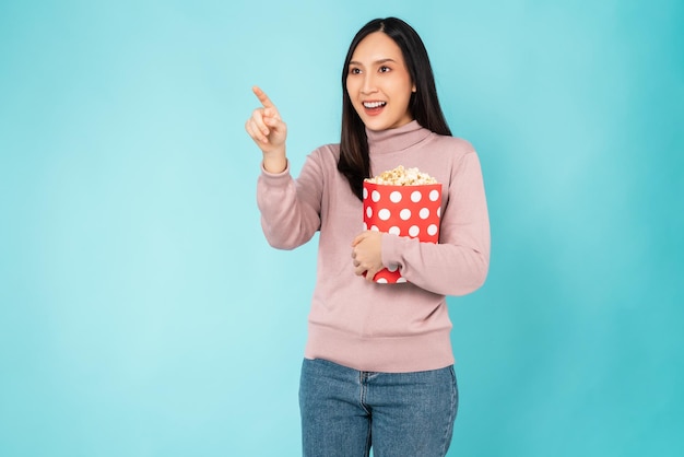 Young woman holding a red bucket of popcorns while pointing front on blue background.