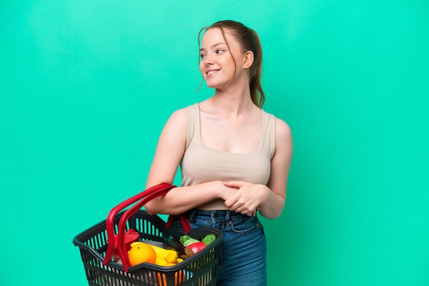 Young woman holding a shopping basket full of food isolated on green background