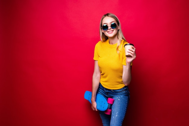 Young woman holding skateboard on shoulder and coffee isolated against red wall