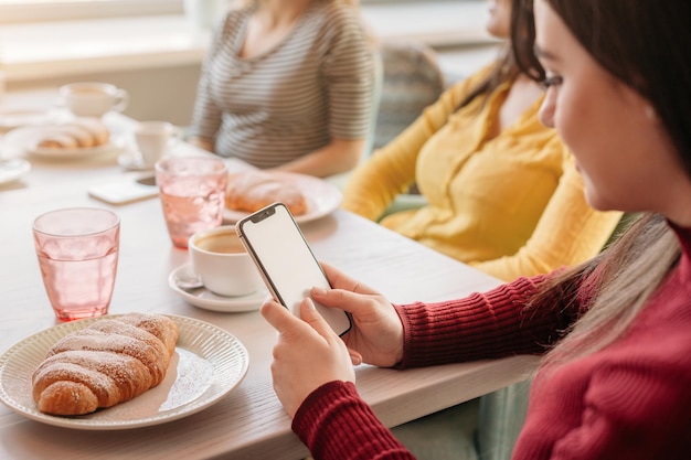 Young woman holding smartphone with blank screen while sitting in cafe with girlfriends