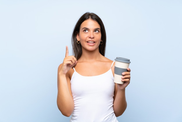 Young woman holding a take away coffee over isolated blue wall intending to realizes the solution while lifting a finger up