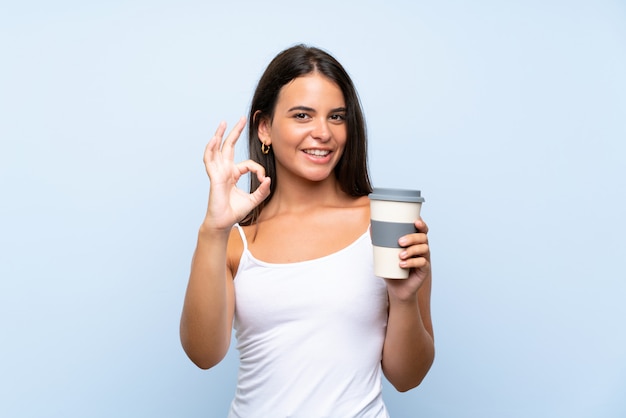 Young woman holding a take away coffee over isolated blue wall showing ok sign with fingers