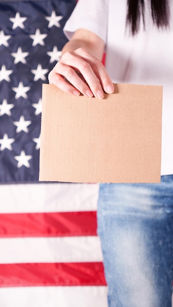 Young woman holds empty cardboard with Space for Text sign against American flag