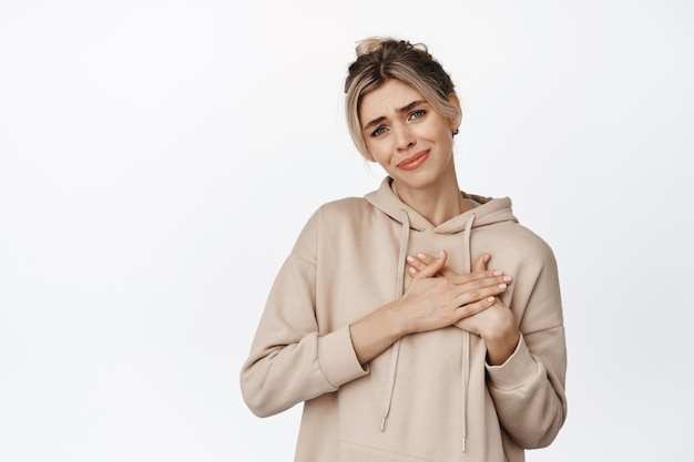 Young woman holds hands on heart and looking touched, heartfelt and emotional about something beautiful, standing on white