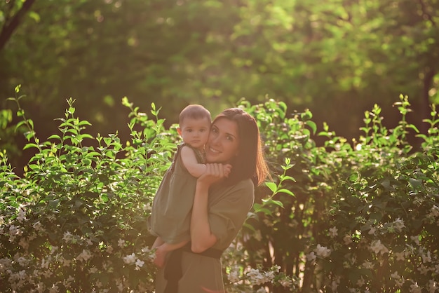 A young woman holds a little baby in her arms. A beautiful mother walks with her daughter in a green park near the jasmine bushes