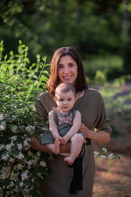 A young woman holds a little baby in her arms. A beautiful mother walks with her daughter in a green park near the jasmine bushes