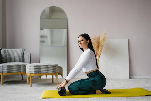 Young woman holds a roller for myofascial release in her hands sitting at home on a gym mat