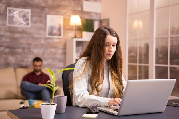 Young woman in home office working on laptop and boyfriend using phone while relaxing on sofa.