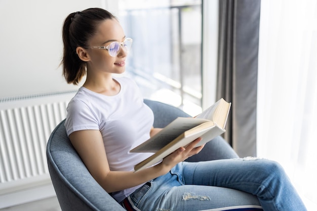 Young woman at home sitting on modern chair in front of window relaxing in her living room reading book