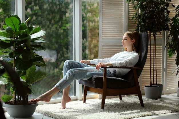 Young woman at home sitting on modern chair near window relaxing in living room