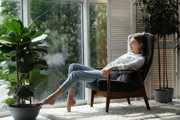 Young woman at home sitting on modern chair near window relaxing in living room