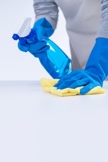 Young woman housekeeper in apron is cleaning wiping down table surface with blue gloves wet yellow rag spraying bottle cleaner closeup design concept