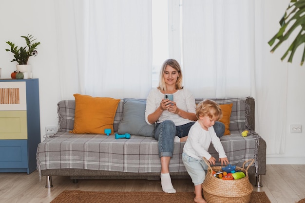 Young woman housewife messaging on phone while sitting on the couch with her little son at home