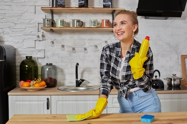 Young woman or housewife in rubber gloves wiping table with microfiber cloth at home kitchen.