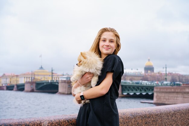 Young woman hugs spitz dog outdoors on embankment of city river in black clothes with long hair happ...