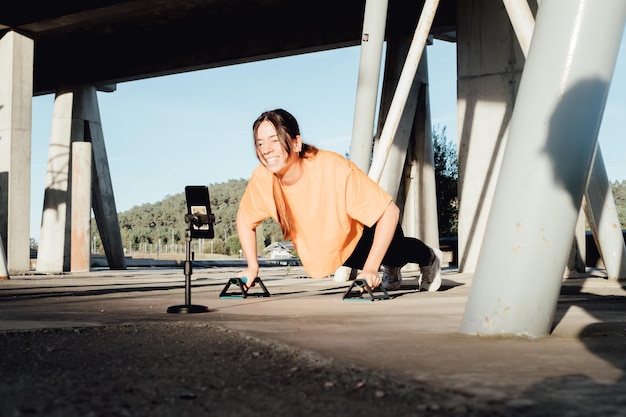 Young woman influencer blogger recording a live streaming on social media with mobile phoneworking up with push ups bar crossTeachingexplaining and doing a demonstrationTraining cheap from home